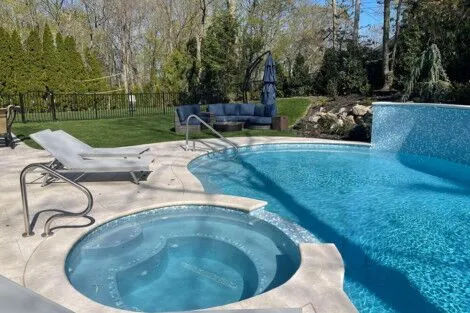 Luxury outdoor pool area with hot tub, lounge chairs, and lush landscaping in Long Island. Beautiful inground pool with hot tub and outdoor furnishings in Long Island.