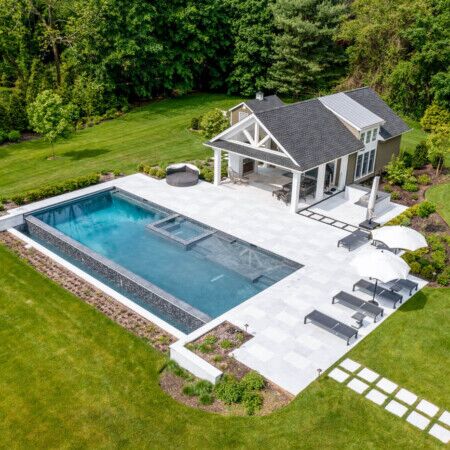 Elegant inground pool with outdoor seating and a pool house in a lush, landscaped backyard. Luxury inground pool with outdoor furnishings in a landscaped backyard.