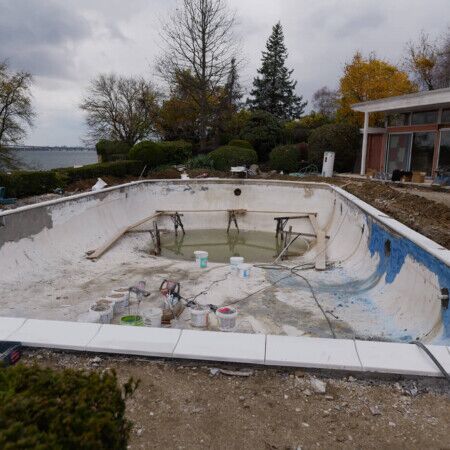 An inground pool undergoing renovation with surrounding landscaping and a modern house in the background. Empty inground pool under renovation with tools and debris present.