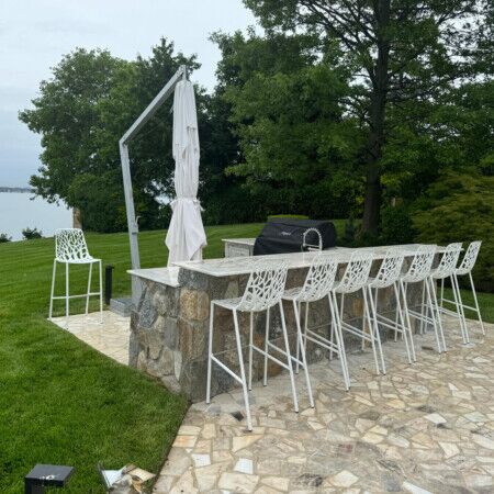 Outdoor bar area with stone counter, bar stools, and umbrella overlooking a scenic green landscape. Outdoor bar with stone counter, bar stools, and umbrella in a lush backyard setting.