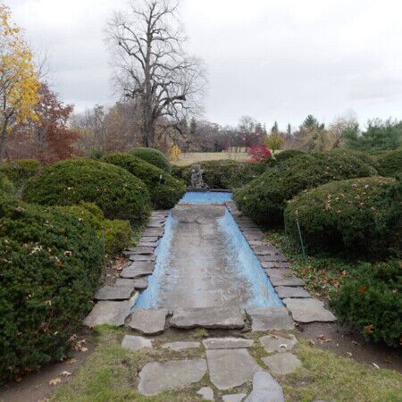 A historic garden fountain with surrounding lush bushes and a stone pathway in Kings Point, reflecting 1950s landscape design. Vintage garden fountain with stone pathway and lush greenery in Kings Point.