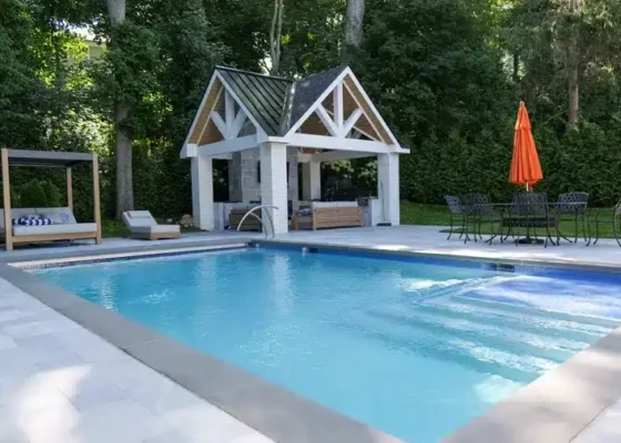 Inground pool with outdoor furniture and gazebo in a lush backyard.