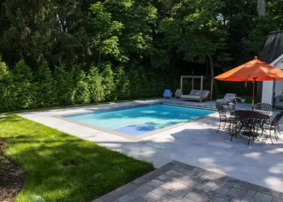 Inground pool with outdoor dining area and umbrella in a lush backyard.