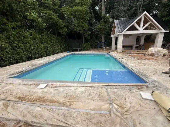 Inground swimming pool with steps and surrounding stone deck in a backyard.