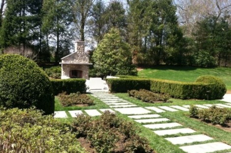 Landscaped garden featuring a stone pathway leading to an outdoor fireplace, surrounded by lush greenery and trimmed bushes. Beautiful landscaped garden with stone pathway and outdoor fireplace.