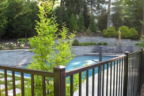Beautiful inground pool with fencing and outdoor furnishings in a lush backyard setting in Long Island. Inground pool with black metal fence and lush greenery in Long Island.
