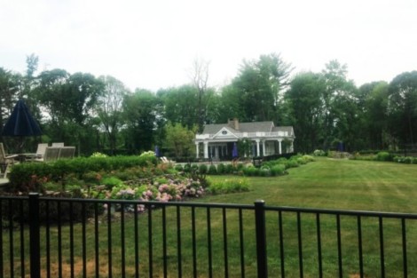 Beautiful backyard with black metal fence, garden, and a large house in the background. Long Island backyard with black metal fence and lush greenery.