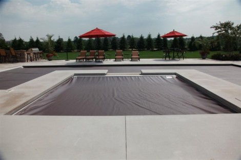 Modern inground pool with a sleek automatic cover, surrounded by outdoor furniture and umbrellas. Inground pool with automatic cover and outdoor seating area.