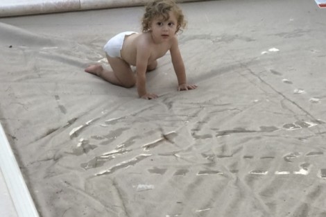 A young child crawling on a pool cover in a backyard with green grass and potted plants. Child crawling on a pool cover in a backyard setting.