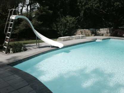 Beautiful inground pool with slide and outdoor furniture in a lush backyard setting. Inground pool with slide and outdoor furnishings in Long Island.