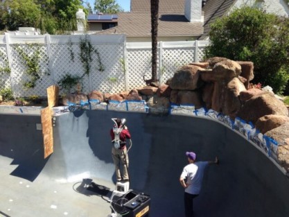 Workers renovating a gunite pool with equipment and natural rock features in the background. Pool renovation with workers applying gunite coating on the pool surface.