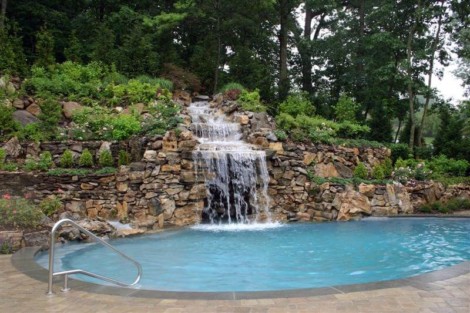 Beautiful inground pool with a waterfall feature surrounded by lush greenery and natural rocks in Long Island. Inground pool with waterfall and natural rock landscaping in Long Island.