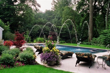 Beautiful inground pool with water fountains surrounded by vibrant garden and outdoor furnishings in Long Island. Inground pool with water fountains and lush garden landscape.