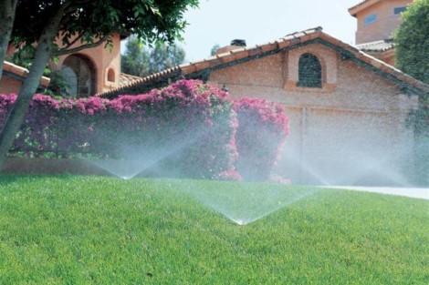 Irrigation system watering a well-maintained lawn in front of a house with outdoor furnishings. Sprinkler watering lush green lawn with house in background.