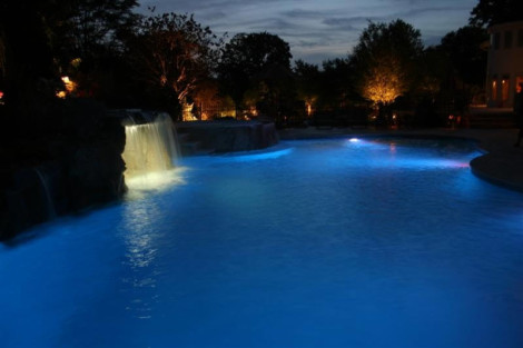 Night view of a beautifully lit inground pool with waterfall and landscape lighting on Long Island. Illuminated inground pool at night with waterfall and landscape lighting.