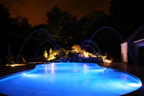 Night view of a beautifully lit inground pool with water features and outdoor lighting on Long Island. Illuminated inground pool at night with water features and outdoor lighting.