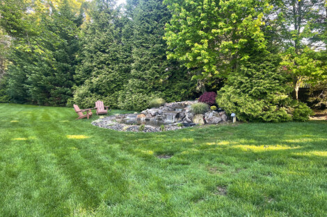Serene garden scene with waterfall, trees, and outdoor chairs for relaxation. Lush garden with waterfall, trees, and outdoor seating area.